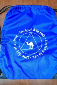 Sac à dos bleu vif, avec un logo contenant un dromadaire et l'inscription trilingue "un jour à la fois".